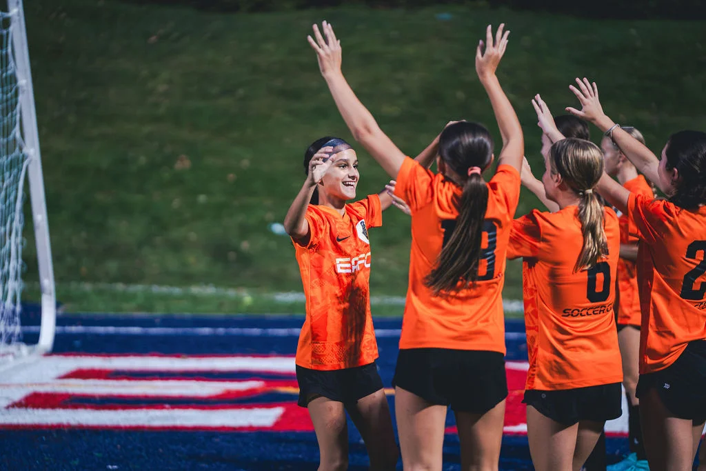 EAFC girls celebrating a goal together in their orange kits.